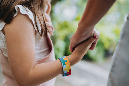 A young girl diagnosed with autism spectrum disorder is wearing an autism awareness bracelet and holding hands with an adult. 