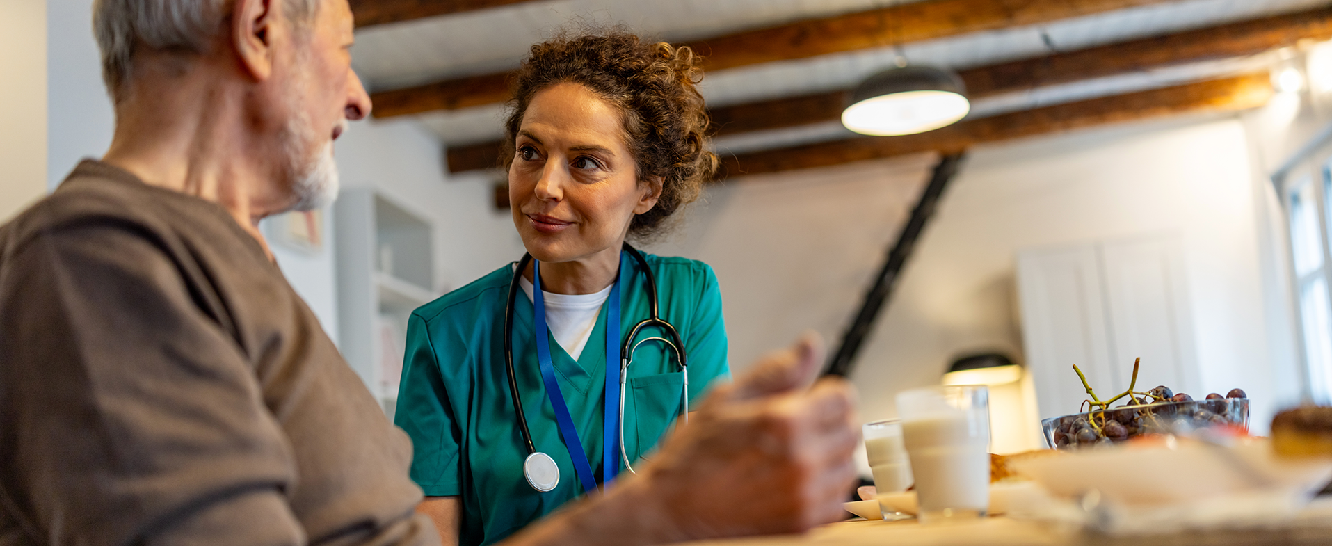 Older man with white hair wearing brown shirt sitting at a table with a medical professional wearing green scrubs. older man talking to medical professional