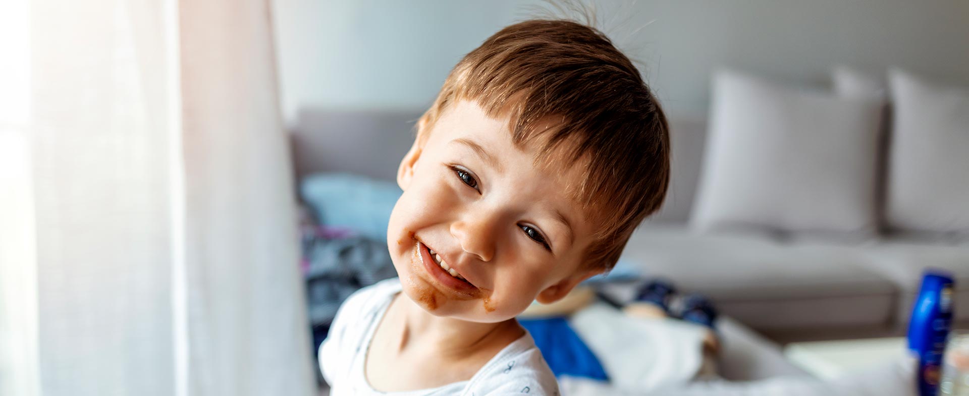 Young boy in a white shirt smiling cutely at the camera. He is holding a piece of toast with smooth peanut butter spead on it and has peanut butter all around his mouth. peanut allergy decline