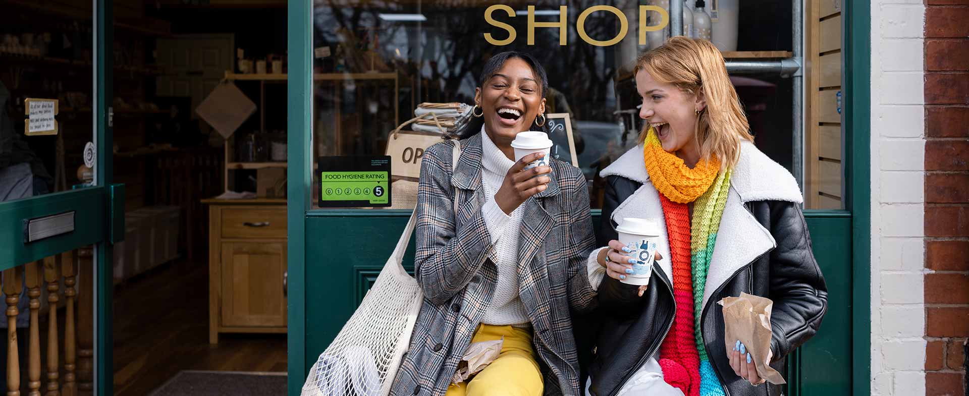 Two women sitting on a bench outside a "Refill Shop," holding coffee cups and smiling. One wears a checked coat and yellow pants, and the other wears a black and white coat with a colorful scarf. The shop door is open, with visible shelves and products inside. rx for laughter