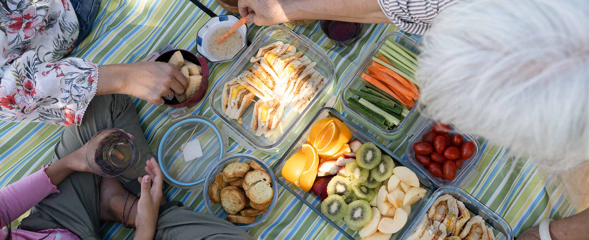 A picnic setup on a striped blanket featuring various foods including sliced fruits, vegetables, sandwiches, crackers, and cheese. Several hands are reaching for the food. wellness on the go