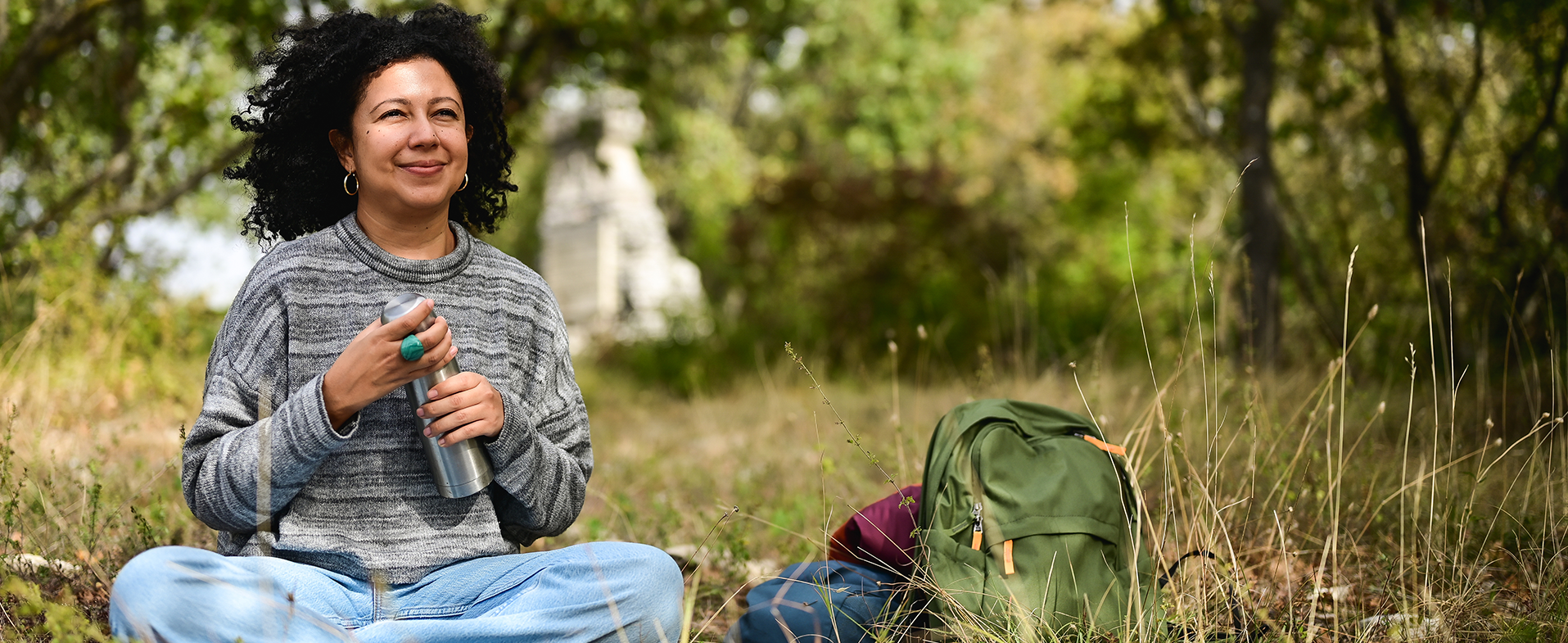 Woman with dark shoulder-length curly hair sitting outside in the grass cross-legged. She is holding a metal water bottle and wearing a gray striped sweater and jeans. dummy desktop Image