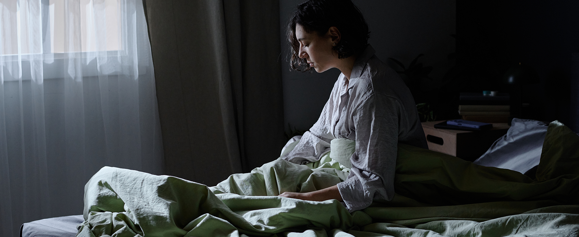 Woman with short dark curly hair sitting up in bed at night. Her eyes are still closed and she is wearing a grayish white pajama shirt. woman awake in bed