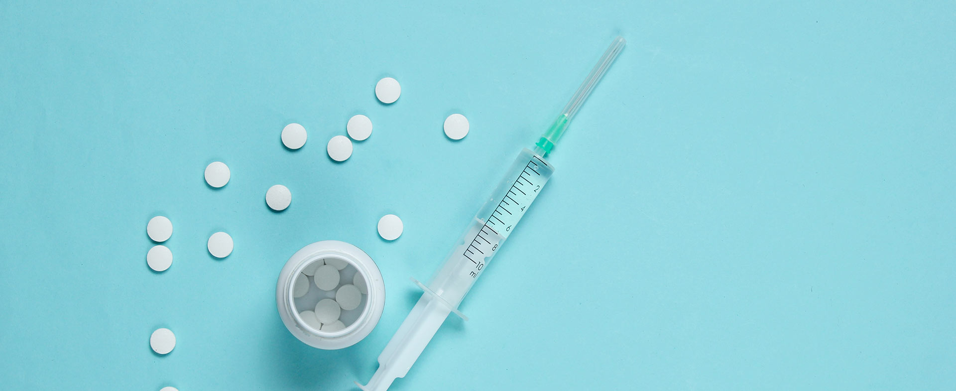 A syringe and a small container with scattered white pills are placed on a light blue surface. allergy shots vs meds