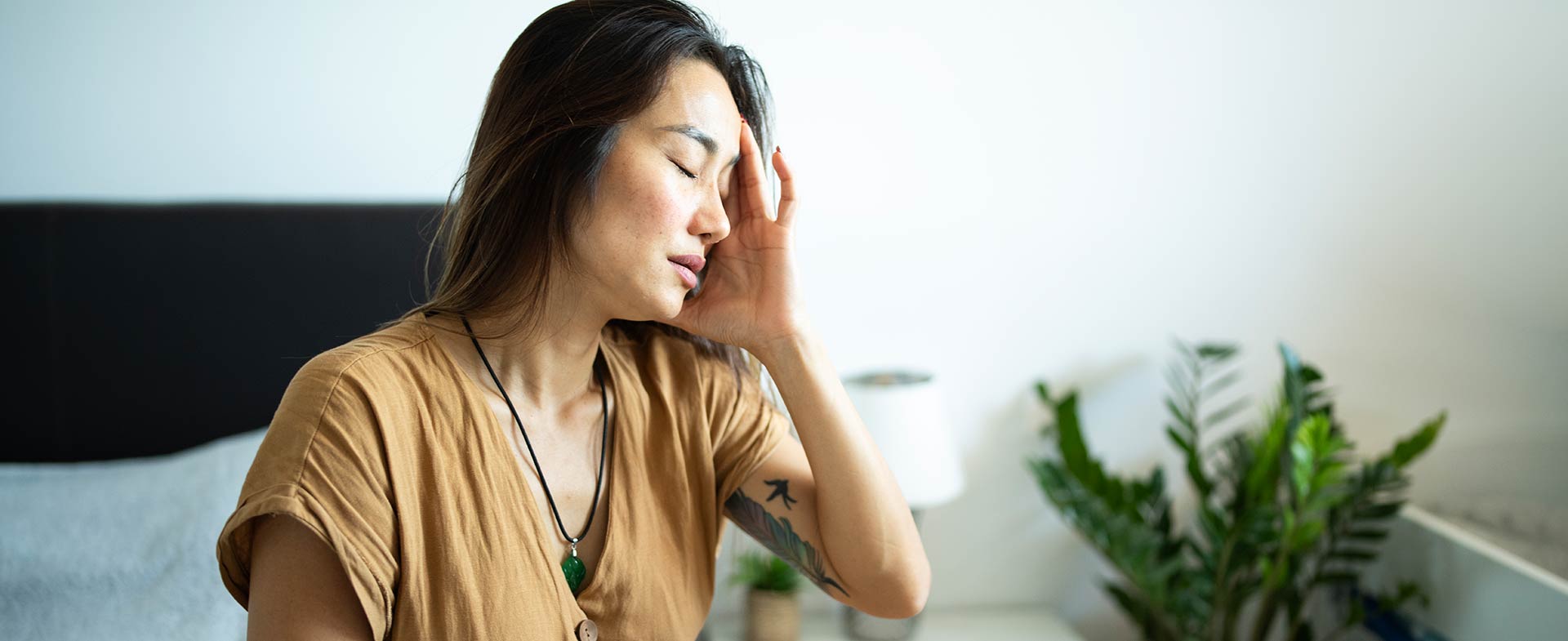 An Asian woman sits on a bed in a room with a white wall, gently touching her head, suggesting discomfort. A plant and a lamp are on a bedside table in the background. cervicogenic headaches