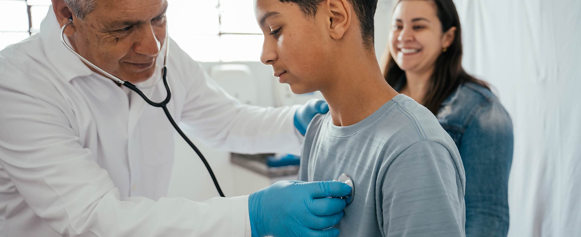 A medical professional uses a stethoscope to listen to a patient's chest during a check-up. Another person is smiling in the background. The setting appears to be a clinic or doctor's office. baseline heart testing for student athletes