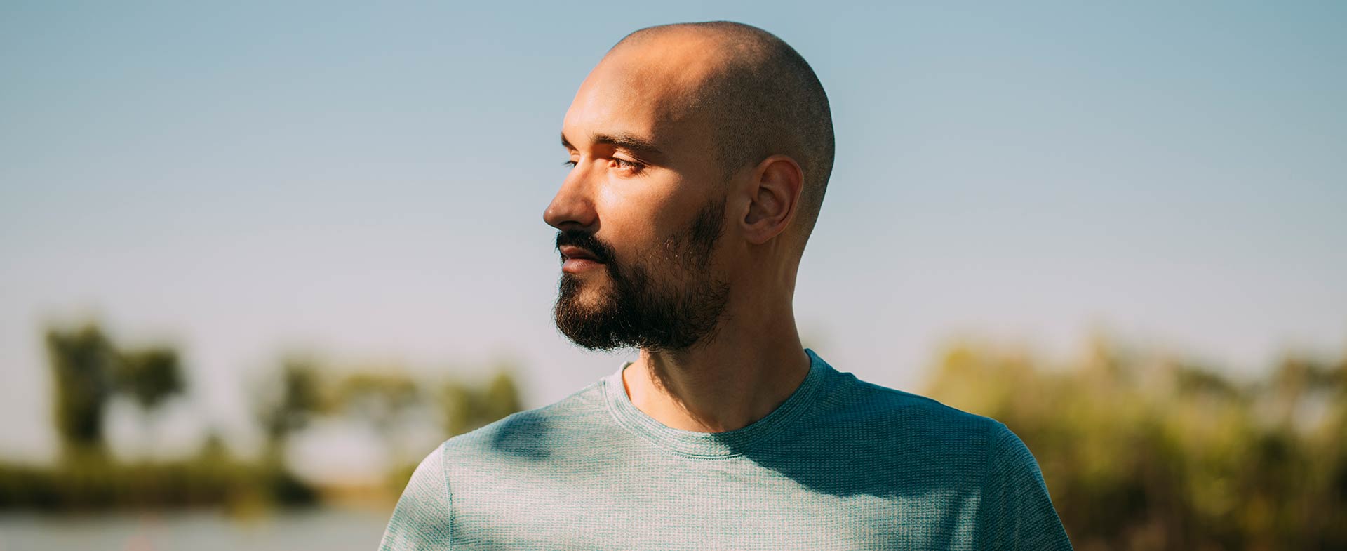 A young bald man with a beard stands outside looking off into the distance. The background shows a blue skies and trees. holistic ways to manage stress
