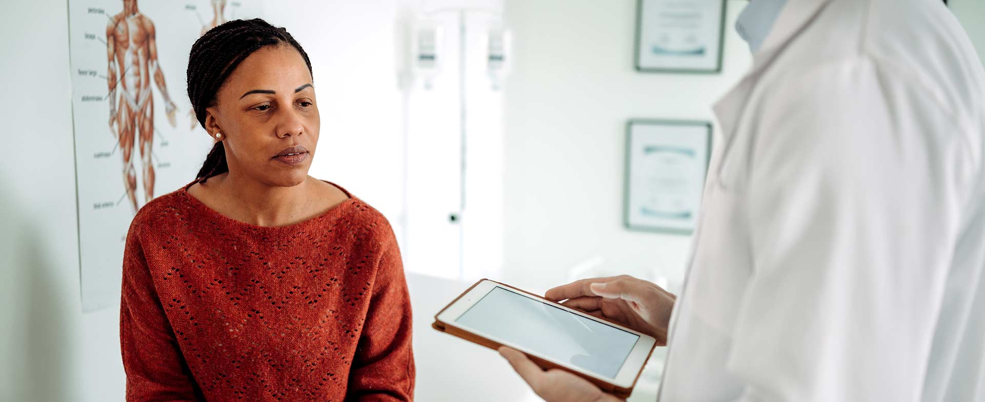 A healthcare professional holding a tablet, speaking with a woman sitting on an examination table. There is a medical diagram on the wall in the background, suggesting a clinical setting. pmos