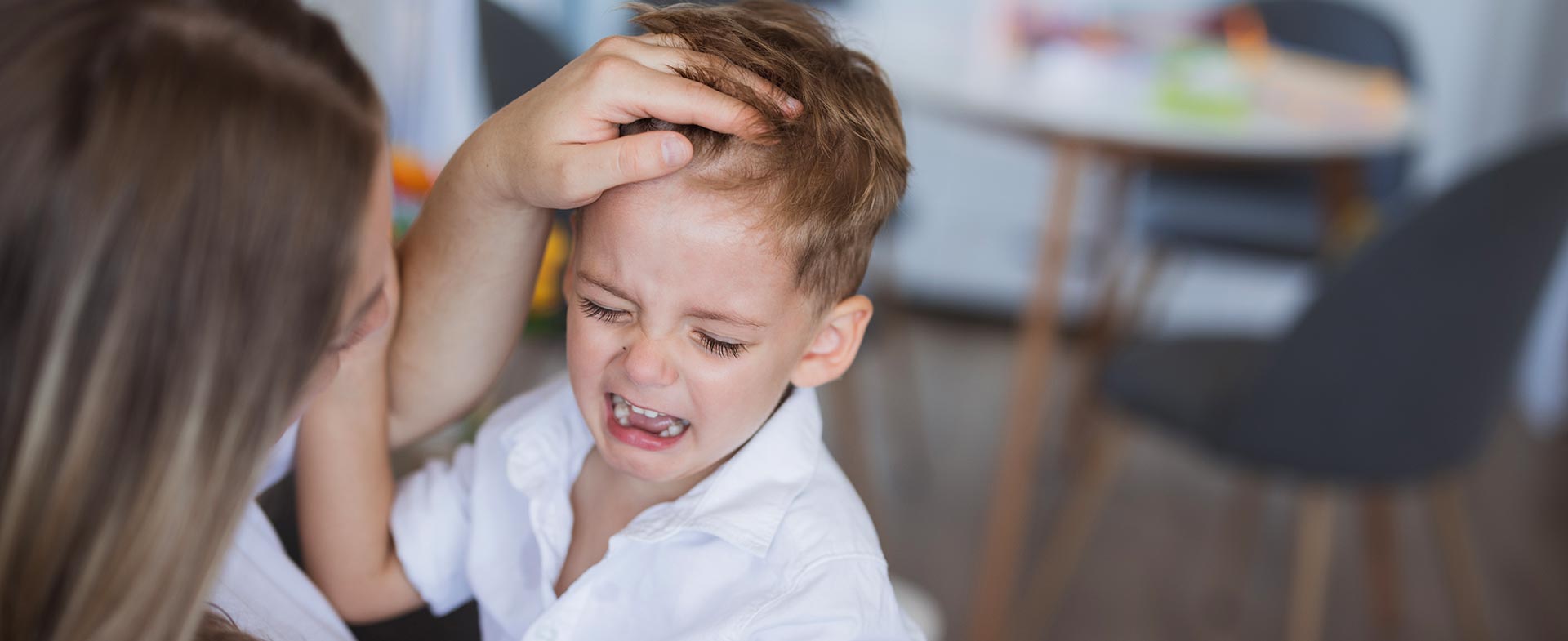 A woman comforting a crying young boy in a home setting. A table and chairs are visible in the background. toddler head bumps