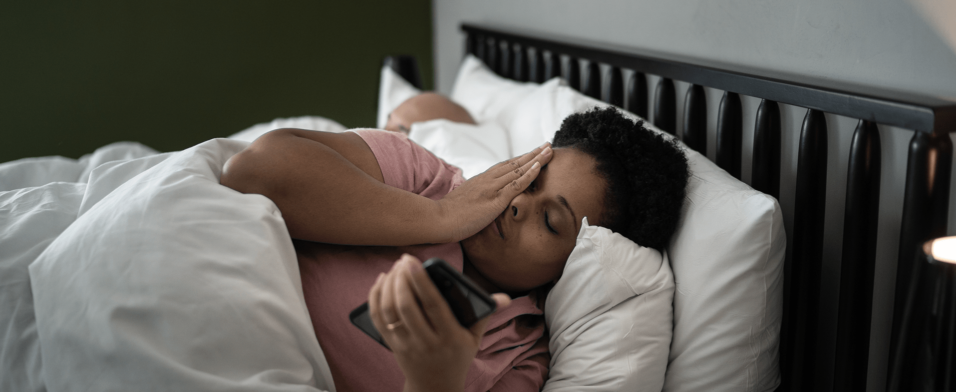 Woman with short curly hair laying in bed wearing a rose-colored t-shirt. She has her hand over her face and is holding her phone. dummy desktop Image