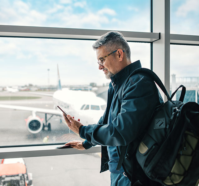 Middle aged man with glasses wearing a navy jacket looking at cell phone in front of an airport window flying and heart heart