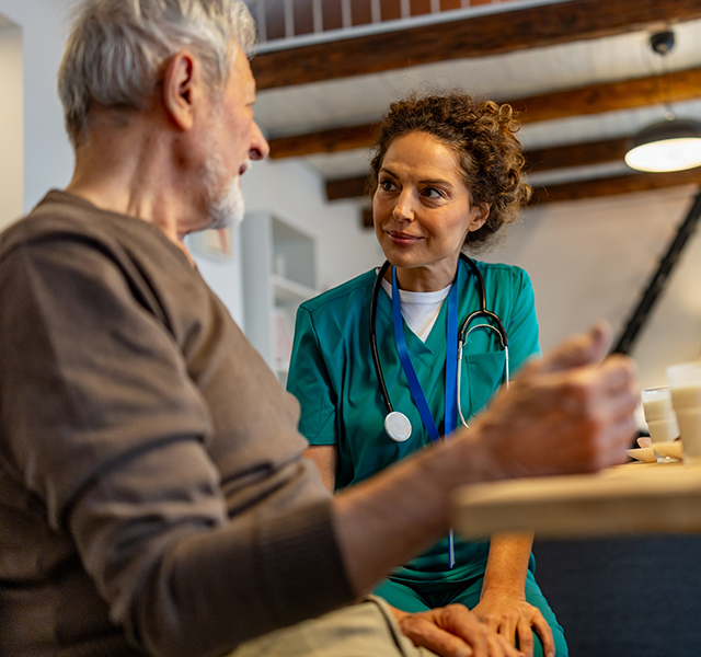 Older man with white hair wearing brown shirt sitting at a table with a medical professional wearing green scrubs. older man talking to medical professional