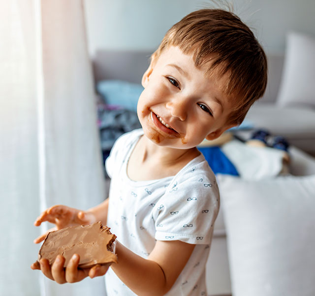 Young boy in a white shirt smiling cutely at the camera. He is holding a piece of toast with smooth peanut butter spead on it and has peanut butter all around his mouth. peanut allergy decline