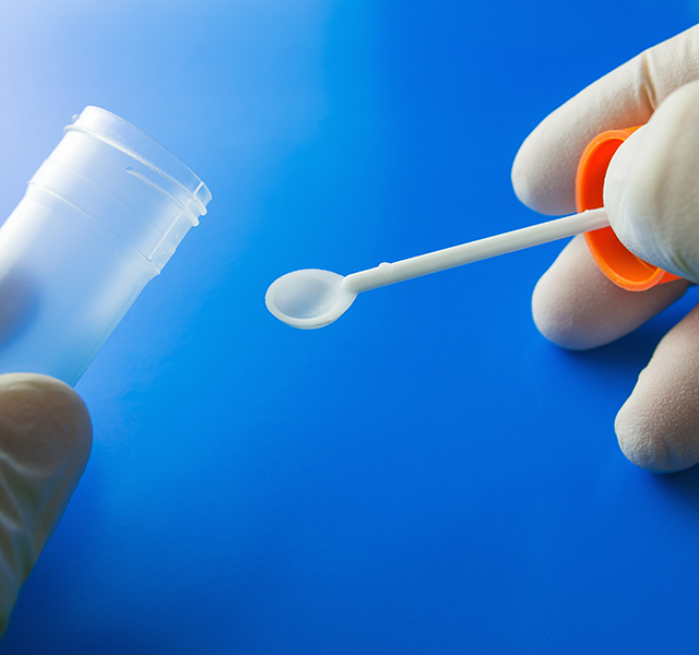 Close-up image of two gloved hands on a blue background holding a clear stool sample collection container. stool collection container