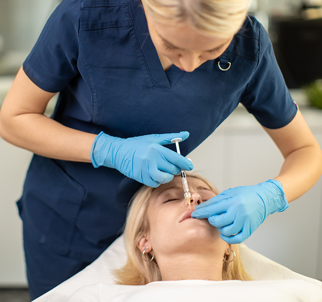 Woman laying back with her eyes closed. She has blonde shoulder-length hair and is getting dermal filler injected into her upper lip. dummy mobile image