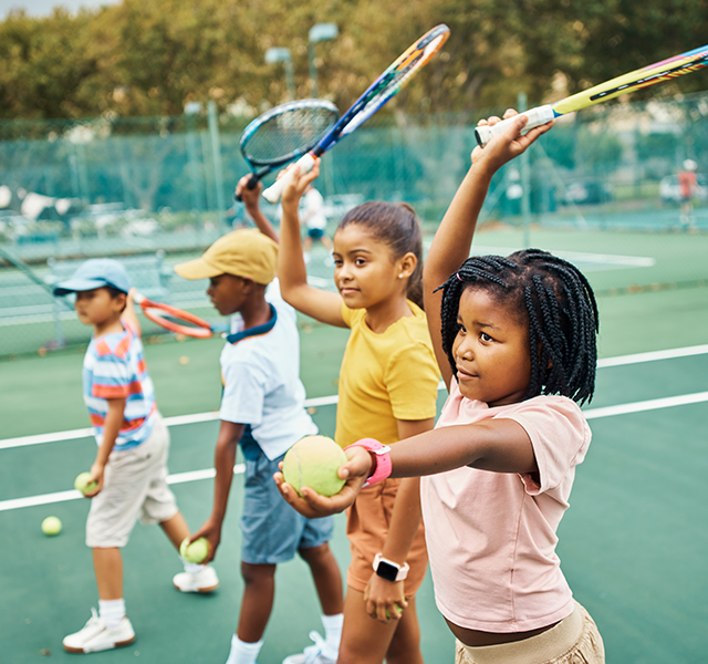 Two girls wearing t-shirts - one light peach and one yellow - and two boys wearing t-shirts - one white with a navy trim and one blue, red and white striped. They are all outside on a tennis court playing tennis. dummy mobile image