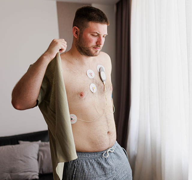 A man with medical electrodes attached to his chest standing near a window in a softly lit room while putting on a button down shirt. The background includes a section of a sofa with cushions. heart monitors