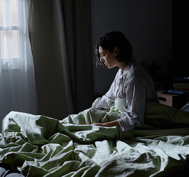 Woman with short dark curly hair sitting up in bed at night. Her eyes are still closed and she is wearing a grayish white pajama shirt. woman sitting up in bed