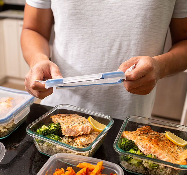 A person in a kitchen is sealing a food container filled with broccoli and salmon. Additional containers with similar meals are on the counter. new dietary guidelines