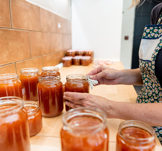 A woman carefully sealing jars of orange jam on a wooden countertop. Multiple filled jars are aligned in the background, and she is wearing an apron with a floral pattern. canning safety