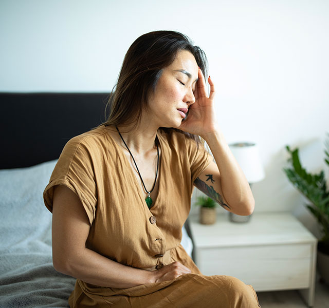 An Asian woman sits on a bed in a room with a white wall, gently touching her head, suggesting discomfort. A plant and a lamp are on a bedside table in the background. cervicogenic headaches