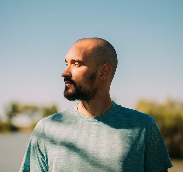 A young bald man with a beard stands outside looking off into the distance. The background shows a blue skies and trees. holistic ways of managing stress