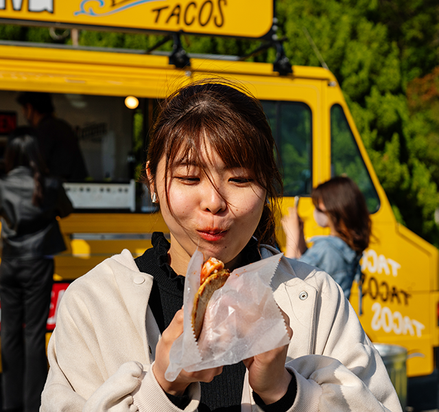 Young woman with dark hair and bangs standing outside in front of a yellow taco truck eating a taco. woman eating taco