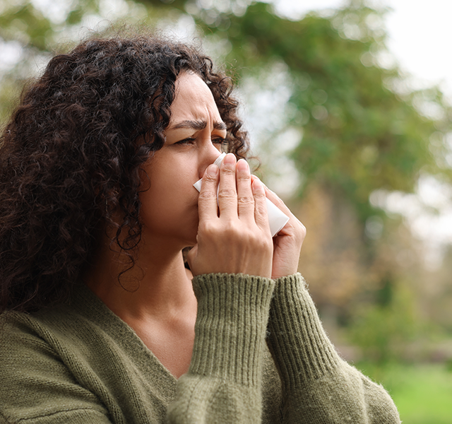 Woman with dark curly hair wearing a green long-sleeve sweater and blowing her nose outside. dummy mobile image