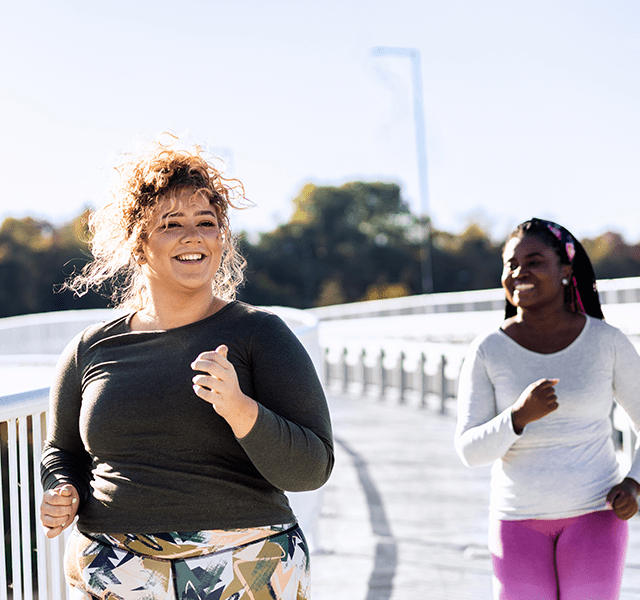 Two friends running outside. One is wearing a black long-sleeved running shirt and multi-colored patterned leggings. The other is wearing a white long-sleeved running shirt and hot pink leggings. Both are smiling. dummy mobile image