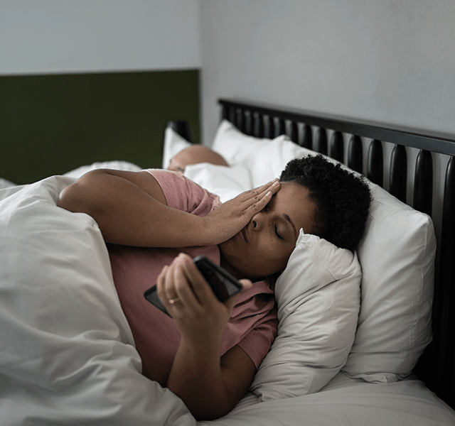 Woman with short curly hair laying in bed wearing a rose-colored t-shirt. She has her hand over her face and is holding her phone. dummy mobile image