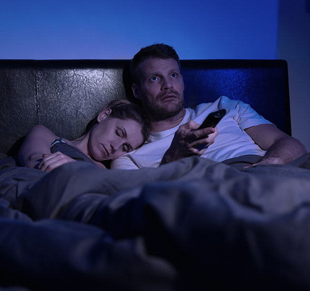 A couple sits in bed at night. The woman is asleep on the man's shoulder while he stays awake and changes the TV channel. early bird vs night owl heart health
